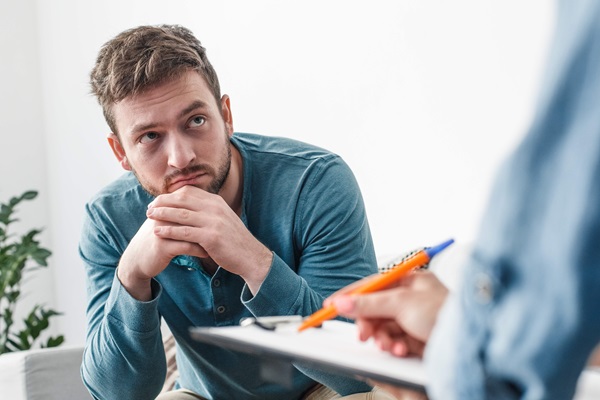 A young male patient holding his chin with a deflated expression looking at a health care provider holding a pen and clipboard in the foreground