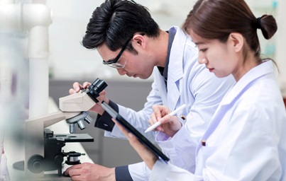 Two researches in lab. One looking into microscope and one writing on tablet.