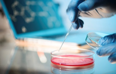 A gloved hand uses a pipette to transfer liquid into a petri dish. A blurred computer screen is visible in the background.